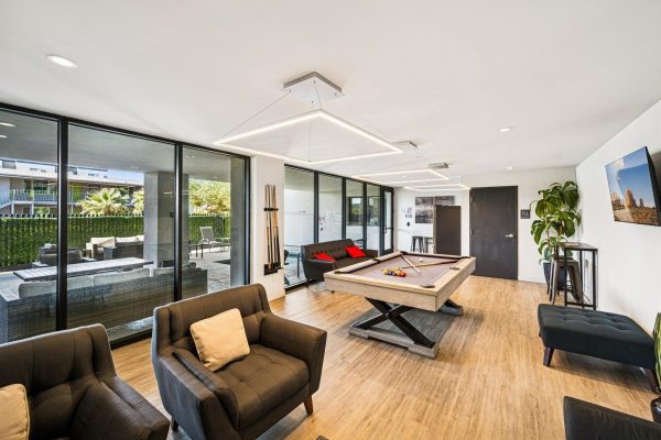 Modern indoor game room with a pool table, black cushioned chairs, and a sofa, plus large glass doors to an outdoor seating area on a sunny patio.